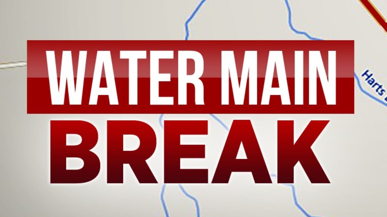 Rochester city street with a water main break, showing water flowing and construction barriers.
